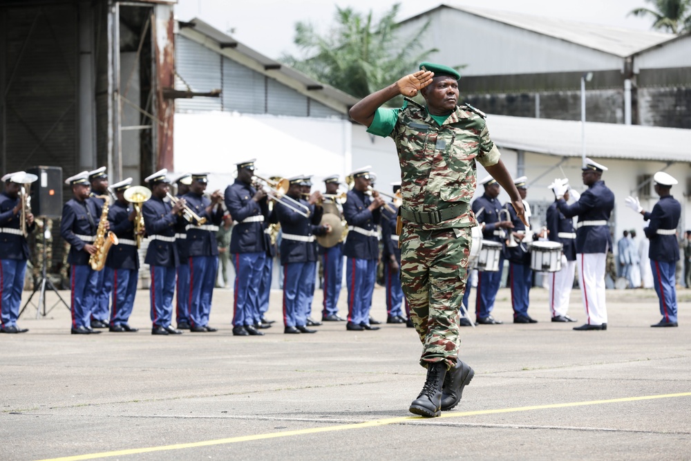 A military officer in camouflage uniform and green beret is saluting while marching across a parade ground. Behind him, a military band in dark blue dress uniforms with red striping is performing, with musicians playing brass instruments, saxophones, and drums. The setting appears to be a formal military ceremony or parade at what looks like a military installation or airfield.