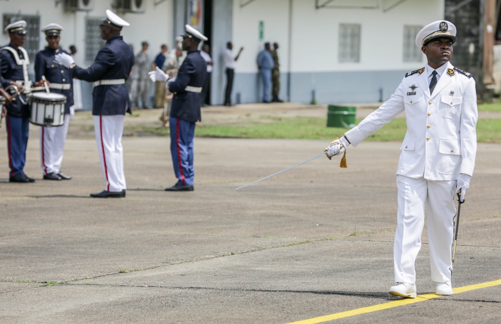 A military ceremony or parade is taking place on what appears to be a parade ground or tarmac. A senior officer in a white dress uniform holds a ceremonial sword in the foreground, while a military band with drummers in blue and red uniforms performs in the background. The setting appears to be an African military installation based on the uniform styles and architectural details.