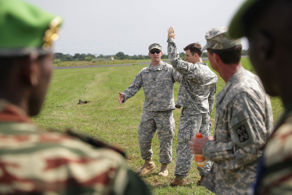 A group of military personnel in different uniforms are gathered outdoors on a grassy field, appearing to be in a briefing or instructional session. Several soldiers in US Army ACU (Army Combat Uniform) pattern camouflage are interacting with personnel wearing green berets and different camouflage patterns, suggesting a multinational military engagement or training exercise. One US soldier has his hand raised, possibly demonstrating or explaining something.