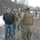 A group of military personnel in digital camouflage uniforms and tactical gear are gathered on a muddy gravel road in a winter environment with snow visible on the ground. A civilian male wearing a black North Face jacket, blue jeans, and sunglasses stands nearby observing or interacting with the uniformed personnel. The setting appears to be an outdoor training exercise or operational site with bare trees and overcast skies in the background.
