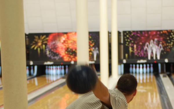 A man is captured mid-release while bowling at a bowling alley, his body leaning forward in a follow-through stance. A dark bowling ball is visible in motion near his raised arm, showing motion blur. Multiple bowling lanes with full pin sets are visible in the background, decorated with colorful fireworks-themed graphics.