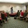 A woman in a black and white patterned dress is presenting or speaking on a small raised platform/stage to an audience seated in banquet-style chairs. The event appears to be a seminar, workshop, or conference held in a hotel or convention center meeting room. The audience consists predominantly of women seated in rows facing the speaker.