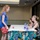 A young woman with red hair and glasses wearing a blue shirt and red shorts stands at a table covered with a tropical floral tablecloth, speaking with two people seated on the other side. The setting appears to be an indoor event space or conference room with recessed lighting. The interaction suggests a registration, check-in, or information table scenario.