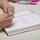 A person is signing a book or large notebook using a pink marker, writing a personalized inscription. The handwriting in pink reads what appears to be 'Sam - Always Fight!!' suggesting this may be a book signing event. Several pink and gray markers are visible in the background on a table.
