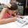 A young girl with a white and yellow flower hair accessory is leaning over a table, writing in an open book with a pink pen. The book appears to contain black-and-white photographs, suggesting it may be a commemorative or autograph book. The setting appears to be an outdoor or semi-outdoor venue with floral-upholstered chairs visible.