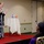 A woman is speaking at a presentation or event in what appears to be a hotel conference room or banquet hall. She is standing on a small raised red-carpeted stage, holding a microphone and gesturing expressively to an audience. At least two audience members are visible in the foreground, seated and facing the speaker.