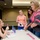A book signing event taking place in what appears to be a hotel ballroom or conference room. A woman seated at a table is signing books and conversing with fans or attendees who are standing in line. Pink markers and water bottles are visible on the signing table.