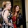 Two women are standing together near what appears to be a bank or ATM vestibule entrance. One woman is wearing a black and white polka dot dress with her hair in a ponytail, while the other is wearing a colorful multicolored dress with a black floral jacket. The woman on the right is smiling broadly, and they appear to be in a friendly interaction.