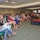 A woman is speaking at a podium on a raised red-carpeted stage in a conference or banquet room, addressing a large seated audience of predominantly women. The event appears to be a seminar, luncheon, or women's conference held in a hotel or country club ballroom. The audience is attentively listening, with rows of chairs filled with attendees extending toward the back of the room.