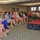 A woman is speaking at a podium on a raised stage to a large audience seated in chairs in what appears to be a hotel conference or banquet room. The audience consists predominantly of women dressed in casual to business-casual attire. The setting suggests a seminar, workshop, or women's networking event.