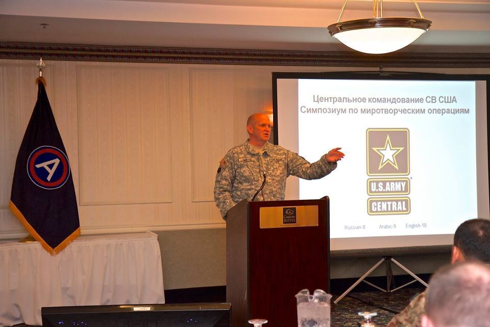 A U.S. Army soldier in ACU (Army Combat Uniform) is presenting at a podium in what appears to be a hotel conference room (Comfort Suites). The presentation slide behind him displays text in Russian reading 'U.S. Army Central Command Peacekeeping Operations Symposium' along with the U.S. Army Central logo. The slide also indicates multilingual support with references to Russian-8, Arabic-9, and English-10.