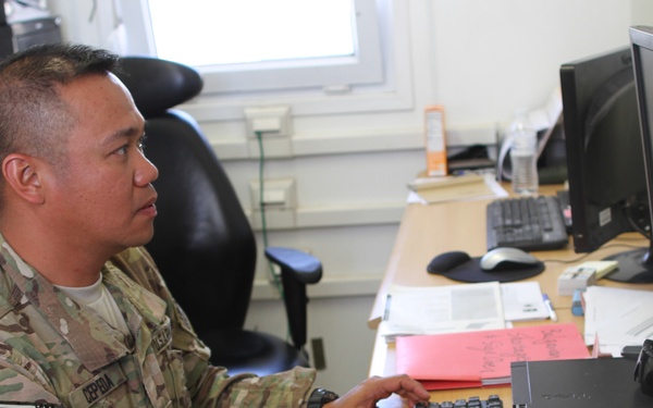 A U.S. Army soldier in MultiCam/OCP camouflage uniform is seated at a desk working on a computer. The soldier's name tape reads 'CEPEDA' and a U.S. flag patch is visible on the left sleeve. The setting appears to be a military administrative office environment.