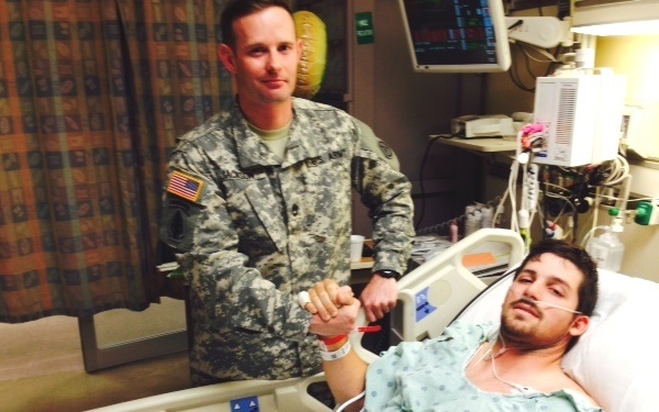 A U.S. Army soldier in ACU (Army Combat Uniform) stands beside a patient lying in a hospital bed. The two individuals are clasping hands in a fist-bump or handshake gesture. The setting is clearly a medical facility with visible monitoring equipment and IV lines attached to the patient.