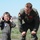 A young child dressed in a miniature military flight suit holds red ear protection headphones while smiling broadly. An adult male in a full military flight suit with patches leans down toward the child, also smiling. The setting appears to be an airfield or military base during daylight hours.