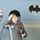 A young child is dressed in a military flight suit, wearing an oversized pilot helmet and holding a oxygen/breathing mask. The child is posed in front of what appears to be a military aircraft fuselage, which features a Batman logo decal and a 9/11 memorial nose art reading 'Never Forget.' The setting appears to be an airshow or aviation museum where children can try on pilot gear.