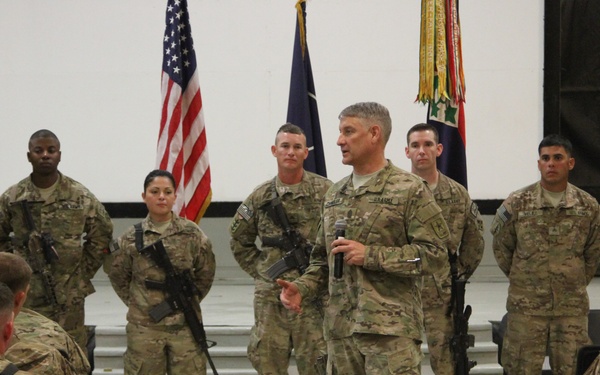 A U.S. Army officer is addressing a formation of soldiers in what appears to be an indoor military briefing or ceremony. The speaker is holding a microphone and standing in front of a group of armed soldiers, with an American flag and military unit colors visible in the background. Seated soldiers in the foreground are listening to the address.