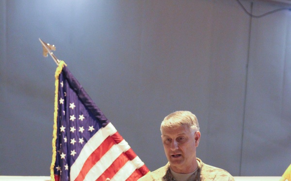A U.S. Army soldier with the name tape 'CHANDLER' is addressing an audience while holding a microphone. He is standing in front of an American flag with gold fringe, appearing to give a speech or briefing to seated personnel. The setting appears to be an indoor military facility or hangar.