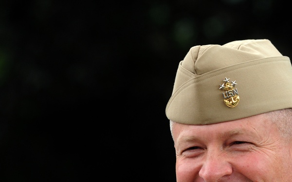 A smiling middle-aged man wearing a United States Navy khaki uniform and garrison cap with a gold USN anchor insignia topped with a silver star. The cap device indicates a senior enlisted rank, consistent with a Fleet Master Chief or Master Chief Petty Officer. The photograph appears to be a professional or semi-formal portrait taken outdoors.