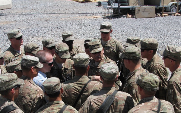 A group of approximately 15-20 military personnel in camouflage uniforms are gathered in a tight circle on a gravel surface at what appears to be a forward operating base or military installation. One individual in civilian clothing (blue shirt) is visible among the uniformed soldiers, suggesting a briefing or meeting with a civilian official or contractor. The setting includes military equipment and structures visible in the background.