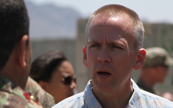 A civilian man in a light blue plaid shirt is engaged in conversation with a person wearing military camouflage uniform, viewed from behind. The setting appears to be an outdoor military installation or base, with mountains visible in the background. Several other military personnel are visible in the background.