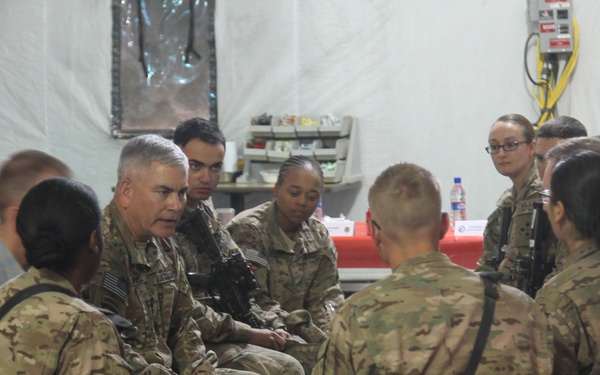 A group of military personnel in MultiCam/OCP camouflage uniforms are gathered in what appears to be a forward operating base or military facility, seated around tables in a discussion or briefing setting. A senior military officer (identifiable by gray hair and rank insignia) appears to be speaking to a group of junior soldiers. The setting suggests an informal leadership engagement or after-action review.