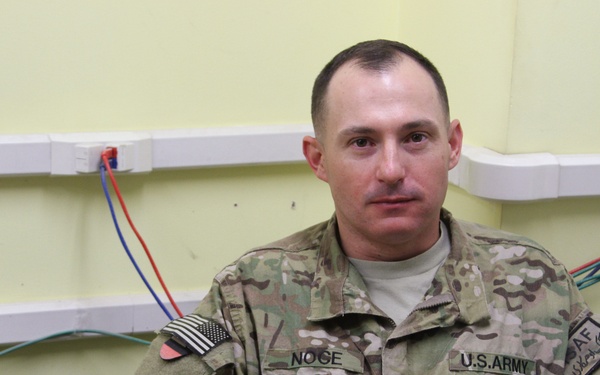 A U.S. Army soldier in MultiCam (OCP) camouflage uniform is seated against a light green wall with exposed colored wiring. His name tape reads 'NOGE' and he wears a U.S. Army tape, an American flag patch on the left shoulder, a ISAF patch on the right shoulder, and a 4th Infantry Division patch. He appears to be in a forward operating base or similar military facility.