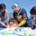 A family of four gathers outdoors around a large decorated sheet cake, with a young girl appearing to be the birthday celebrant. An adult male wearing a US Navy jersey and New York Yankees cap assists the laughing toddler girl in cutting or touching the cake, while a woman in a plaid shirt and an older boy look on. The setting appears to be an outdoor event on a clear, sunny day.