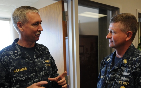 Two U.S. Navy personnel in Navy Working Uniform (NWU) Type I digital camouflage are engaged in conversation in what appears to be an office or administrative building. The individual on the left has the name tape 'ALEXANDER' visible and wears a single star insignia indicating flag officer rank (Rear Admiral Lower Half). The individual on the right wears 'U.S. NAVY' tape and has a submarine warfare qualification badge visible.