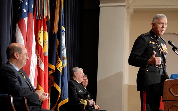 A formal military ceremony or official event is taking place in what appears to be a government or military building. A senior officer in dress uniform is speaking at a wooden podium while two other individuals — one in military dress uniform and one in civilian attire — are seated nearby. Multiple flags, including the U.S. flag, are displayed in the background.