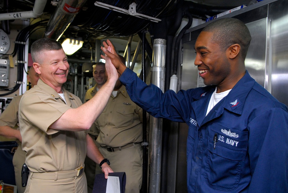 Two U.S. Navy personnel are sharing a high-five gesture aboard what appears to be a naval vessel, both smiling broadly. One individual is wearing khaki service uniform suggesting senior rank, while the other is in blue coveralls labeled 'U.S. Navy'. Additional sailors are visible in the background of the ship's interior corridor.