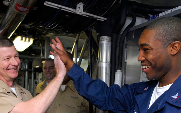 Two U.S. Navy personnel are sharing a high-five gesture aboard what appears to be a naval vessel, both smiling broadly. One individual is wearing khaki service uniform suggesting senior rank, while the other is in blue coveralls labeled 'U.S. Navy'. Additional sailors are visible in the background of the ship's interior corridor.