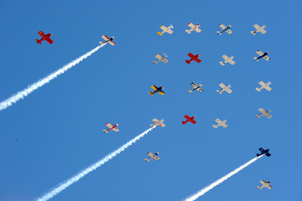 An airshow featuring a large formation of small propeller aircraft flying against a clear blue sky. Multiple aircraft display varied color schemes including red, yellow, white, and blue liveries. At least three aircraft are producing visible white smoke trails, suggesting a coordinated aerobatic display.
