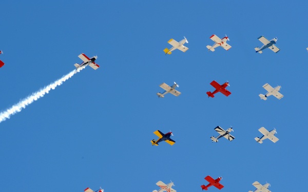 An airshow featuring a large formation of small propeller aircraft flying against a clear blue sky. Multiple aircraft display varied color schemes including red, yellow, white, and blue liveries. At least three aircraft are producing visible white smoke trails, suggesting a coordinated aerobatic display.