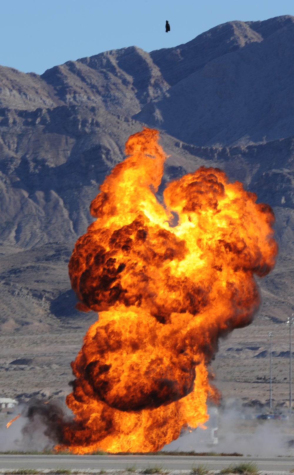 A large fireball explosion rises dramatically against a backdrop of arid desert mountains under a clear blue sky. The explosion appears to be a controlled detonation or pyrotechnic display, likely at an airshow or military demonstration given the flat ground and infrastructure visible at the base. A small dark airborne object is visible near the top of the frame above the mountain ridgeline.