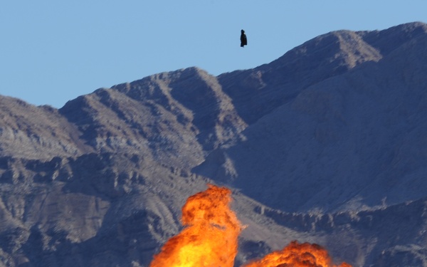A large fireball explosion rises dramatically against a backdrop of arid desert mountains under a clear blue sky. The explosion appears to be a controlled detonation or pyrotechnic display, likely at an airshow or military demonstration given the flat ground and infrastructure visible at the base. A small dark airborne object is visible near the top of the frame above the mountain ridgeline.