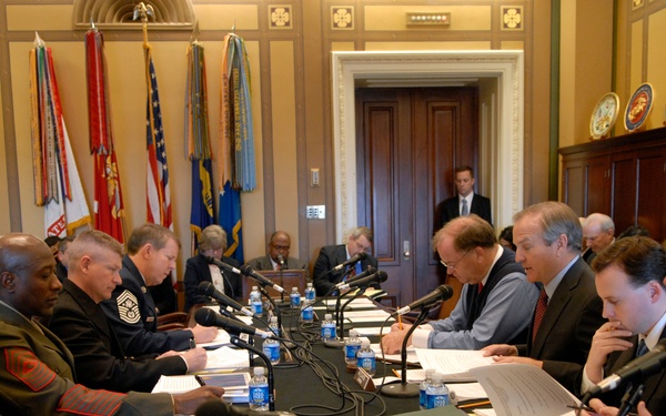 A formal congressional or governmental hearing is taking place in an ornate official chamber, with military personnel in dress uniforms seated on one side of a long conference table facing civilian officials on the other side. Multiple flags representing U.S. military branches are displayed prominently in the background. The setting appears to be a U.S. Capitol or federal building committee room.