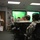 A U.S. Army soldier is conducting a briefing or training session for a group of fellow soldiers seated in office chairs in what appears to be a military classroom or conference room. The presenter, standing to the left near a whiteboard/flipchart, is holding a marker and papers while addressing the group. Multiple uniformed personnel in ACU (Army Combat Uniform) pattern camouflage are attentively listening.