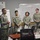 A group of U.S. Army soldiers in ACU (Army Combat Uniform) camouflage are participating in a military training or briefing session. One soldier on the left appears to be presenting or speaking to the group, while four others stand attentively in the background. The setting is a classroom or conference room associated with the 4960th MFTB (Military Force Training Battalion).
