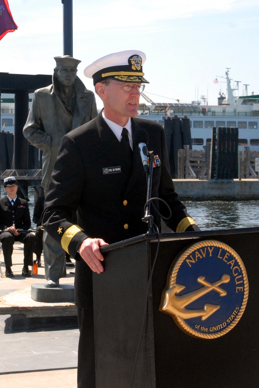 A senior U.S. Navy officer in dress uniform is speaking at a Navy League of the United States podium at an outdoor waterfront ceremony. Behind him stands a bronze statue of a sailor, and a vessel is visible in the harbor background. Additional uniformed personnel are seated to the left.