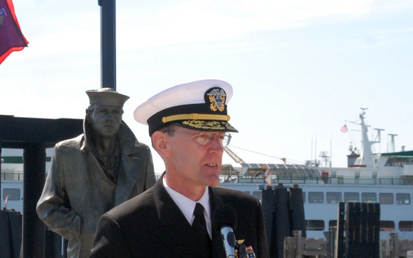 A senior U.S. Navy officer in dress uniform is speaking at a Navy League of the United States podium at an outdoor waterfront ceremony. Behind him stands a bronze statue of a sailor, and a vessel is visible in the harbor background. Additional uniformed personnel are seated to the left.