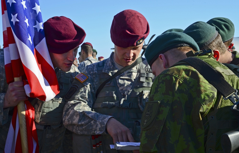 The image shows a group of military personnel from at least two different nations interacting outdoors. U.S. Army soldiers wearing maroon berets and ACU (Army Combat Uniform) pattern camouflage are engaging with soldiers wearing dark green berets and woodland camouflage uniforms. One soldier is holding a U.S. flag while others appear to be reviewing a document together.