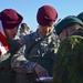 The image shows a group of military personnel from at least two different nations interacting outdoors. U.S. Army soldiers wearing maroon berets and ACU (Army Combat Uniform) pattern camouflage are engaging with soldiers wearing dark green berets and woodland camouflage uniforms. One soldier is holding a U.S. flag while others appear to be reviewing a document together.