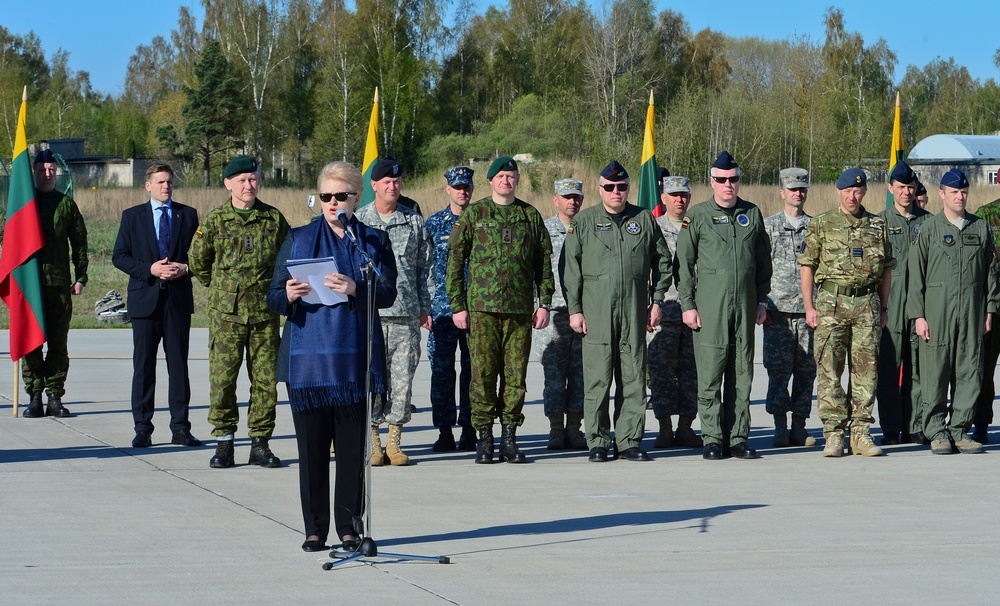 A civilian woman in a blue shawl is delivering a speech at a podium microphone during what appears to be a military ceremony. Behind her stands a formation of military personnel in various uniforms alongside a civilian in a suit, with Lithuanian flags (yellow, green, red tricolor) visible in the background. The event appears to take place on a military airfield or parade ground.
