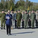 A civilian woman in a blue shawl is delivering a speech at a podium microphone during what appears to be a military ceremony. Behind her stands a formation of military personnel in various uniforms alongside a civilian in a suit, with Lithuanian flags (yellow, green, red tricolor) visible in the background. The event appears to take place on a military airfield or parade ground.