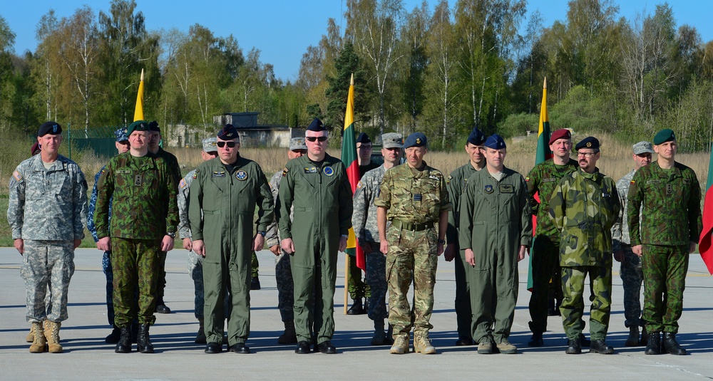 A group of military personnel from multiple nations are standing in formation on a tarmac or parade ground during what appears to be a multinational military ceremony or exercise. Several Lithuanian flags (yellow, green, and red tricolor) are visible in the background, suggesting the event is taking place in or involves Lithuania. Personnel are wearing a variety of uniforms including US Army ACUs, flight suits, British MTP camouflage, and Lithuanian Armed Forces uniforms.