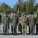 A group of military personnel from multiple nations are standing in formation on a tarmac or parade ground during what appears to be a multinational military ceremony or exercise. Several Lithuanian flags (yellow, green, and red tricolor) are visible in the background, suggesting the event is taking place in or involves Lithuania. Personnel are wearing a variety of uniforms including US Army ACUs, flight suits, British MTP camouflage, and Lithuanian Armed Forces uniforms.