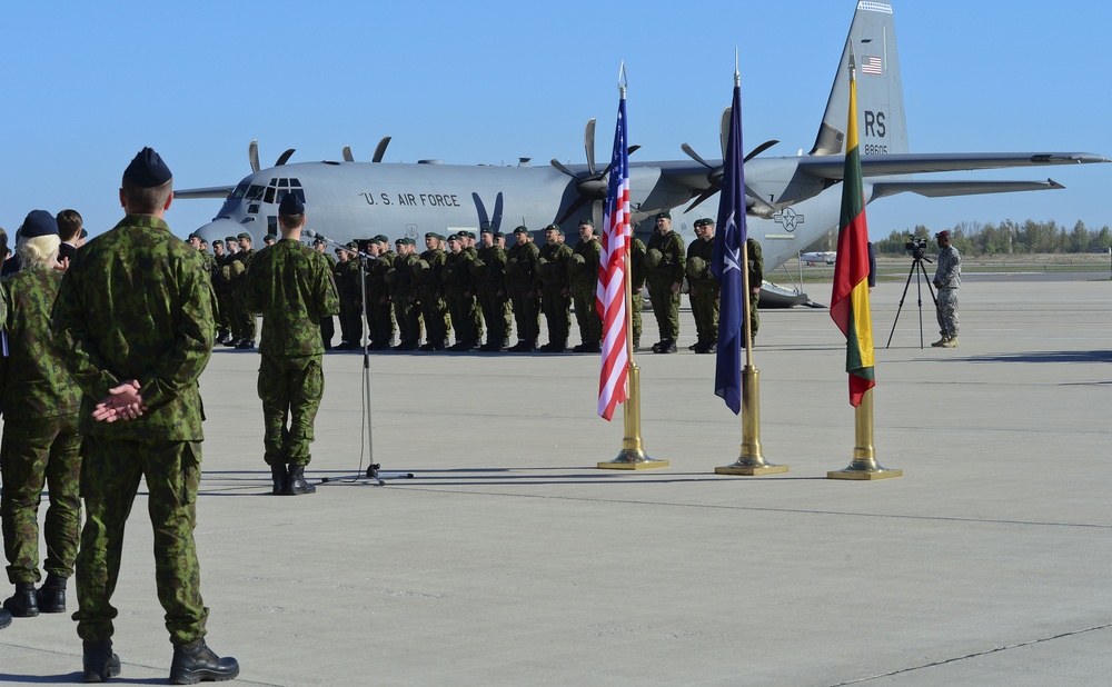 A military ceremony is taking place on an airfield tarmac with troops standing in formation. A U.S. Air Force C-130 Hercules transport aircraft (tail number 88605, unit code RS) serves as the backdrop. Three flags are displayed — the United States flag, a NATO flag, and what appears to be the Lithuanian flag — suggesting a multinational military event.
