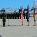 A military ceremony is taking place on an airfield tarmac with troops standing in formation. A U.S. Air Force C-130 Hercules transport aircraft (tail number 88605, unit code RS) serves as the backdrop. Three flags are displayed — the United States flag, a NATO flag, and what appears to be the Lithuanian flag — suggesting a multinational military event.
