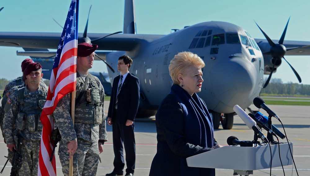 A formal military ceremony or press event taking place on an airport tarmac, featuring a woman speaking at a podium with multiple microphones. U.S. Army soldiers in combat uniforms stand at attention holding an American flag, while a civilian in a suit stands nearby. A large military transport aircraft, consistent with a C-130 Hercules marked with U.S. Air Force markings, is prominently visible in the background.