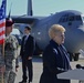 A formal military ceremony or press event taking place on an airport tarmac, featuring a woman speaking at a podium with multiple microphones. U.S. Army soldiers in combat uniforms stand at attention holding an American flag, while a civilian in a suit stands nearby. A large military transport aircraft, consistent with a C-130 Hercules marked with U.S. Air Force markings, is prominently visible in the background.